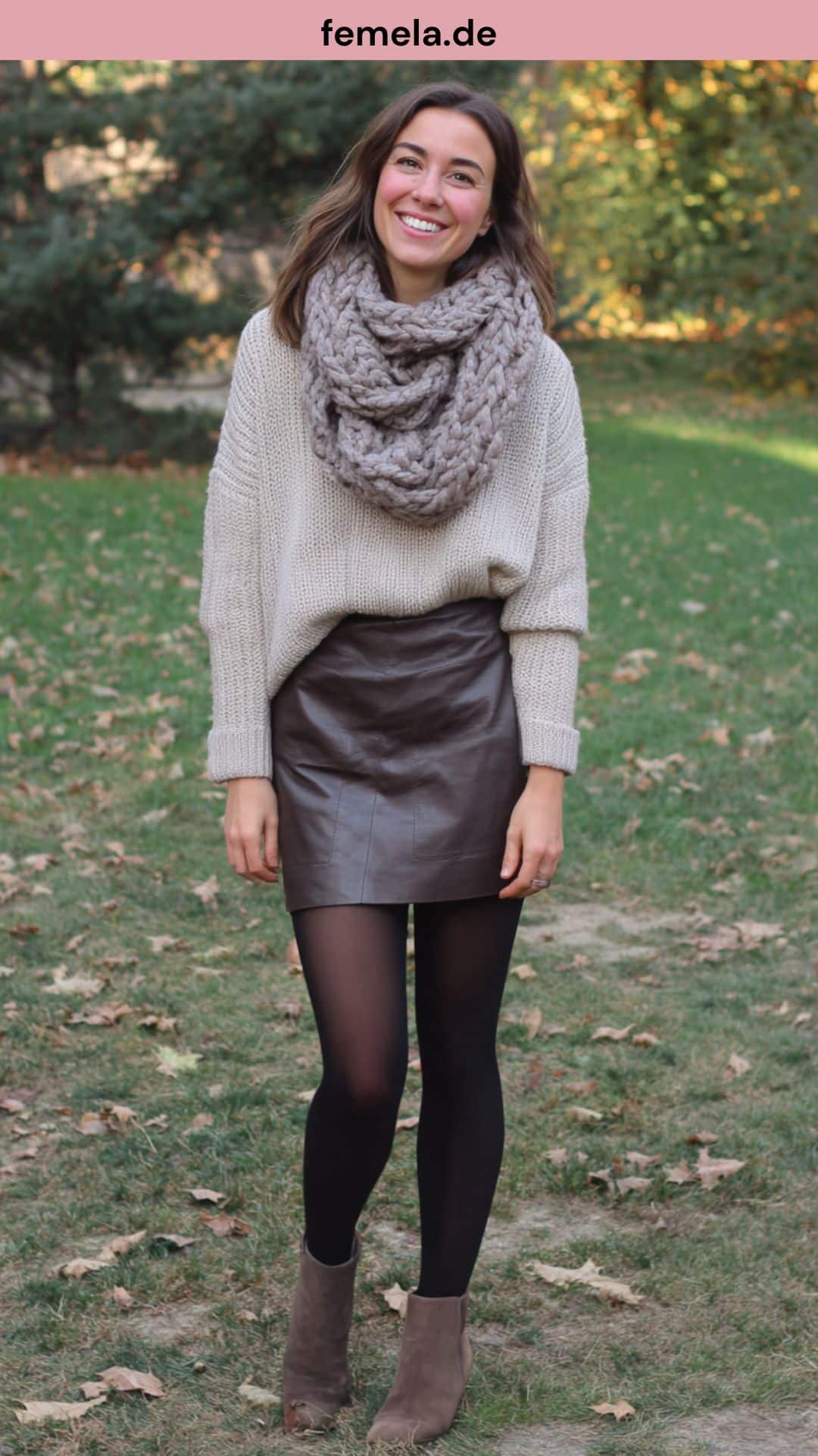 Eine Frau in einem kuscheligen Winter-Outfit mit braunem A-Linien-Lederrock, einem beigefarbenen Oversized-Strickpullover und Stiefeln.